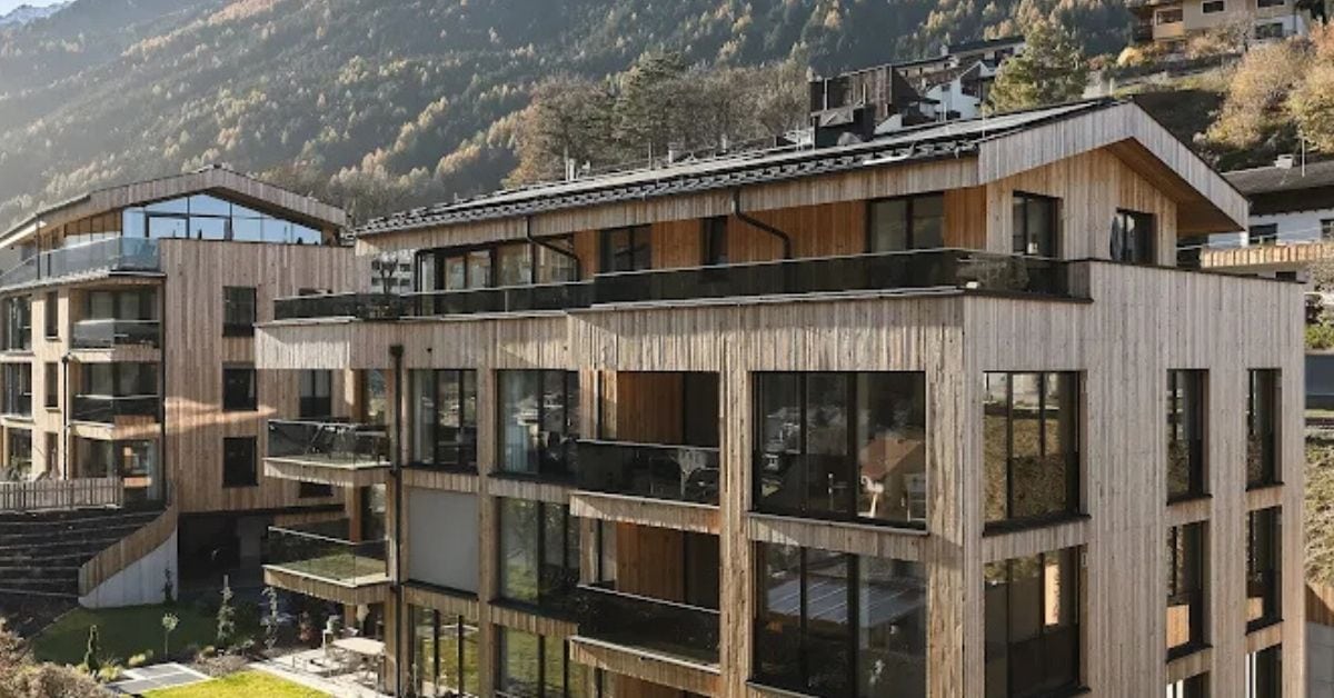 Modern wooden apartment buildings with large windows and balconies are set against a backdrop of wooded hills and mountains in the sunny, scenic Stubai region.